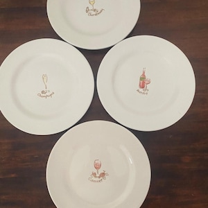 May include: A set of four white ceramic plates, each featuring a unique illustration of a wine glass or bottle and a specific wine name. The designs include a Chardonnay glass, a Champagne flute, a Merlot bottle with glass, and a Cabernet glass with grapes.