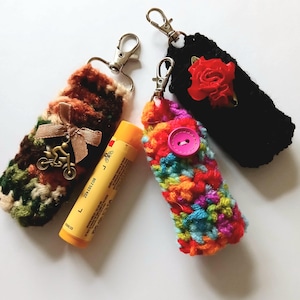 Pode incluir: Três porta-bálsamos labiais de crochê com chaveiros. O primeiro é marrom, verde e bege com um pequeno amuleto de motocicleta e um laço marrom. O segundo é preto com um enfeite de rosa vermelha. O terceiro é multicolorido com um botão rosa.