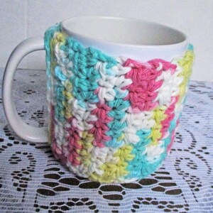 May include: A white ceramic mug with a crocheted cozy in pink, yellow, blue, and white. The cozy is made with a simple stitch pattern and has a geometric design.
