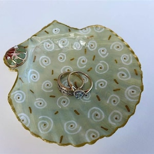 May include: A decorative scallop shell dish painted in pale green with white spiral patterns and gold accents along the scalloped edges. A small, colourful shell embellishment is attached to the top left. Two gold rings rest in the centre.