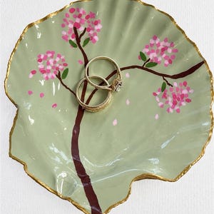 May include: A sage green scallop-shaped trinket dish with a hand-painted brown tree branch featuring pink blossoms and small green leaves. The edges of the dish are trimmed in gold paint. Two gold-toned metal rings rest in the centre of the dish.
