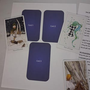 May include: A tarot card reading layout featuring three blank purple rectangular placeholders labeled Card 1, Card 2, and Card 3. The layout includes the Two of Swords, Ace of Cups, and Four of Swords tarot cards, alongside a printed interpretation sheet.