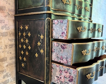 May include: A tall, antique-style dresser with a dark green and black paint finish. The dresser has ornate gold accents and decorative details. The drawers are lined with floral patterned paper.
