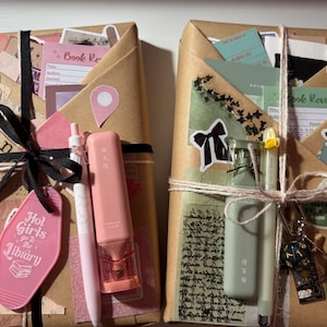May include: Two rectangular packages wrapped in brown paper, decorated with stickers, ribbons, and stationery. The left package features a black ribbon, a pink pen, a pink highlighter, and a pink keychain. The right package has twine, a green pen, a green highlighter, and a multi-sided die keychain.
