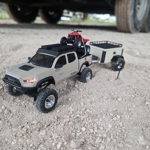 Könnte beinhalten: Ein beigefarbenes Toyota-Pickup-Modell mit schwarzer Verkleidung, einem Dachträger mit einem kleinen roten Quad und einem passenden beigen Anhänger am Heck. Das Fahrzeug verfügt über Geländereifen, silberne Felgen und sichtbare Markenzeichen.