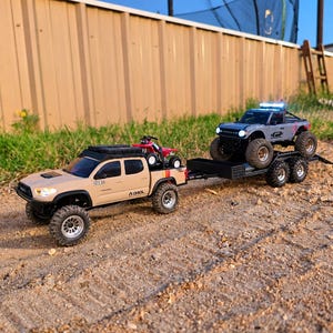 Può includere: Un pickup radiocomandato color sabbia con portapacchi e un piccolo quad rosso nel cassone. Il pickup traina un rimorchio nero a pianale che trasporta un SUV radiocomandato grigio con luci blu sul tetto accese e grandi pneumatici fuoristrada.