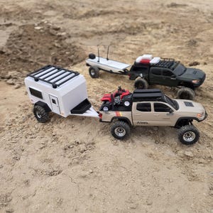 May include: A tan pickup truck with a roof rack and a red toy ATV in the bed, towing a white camper trailer. In the background, a dark gray pickup truck with a roof rack tows a flatbed trailer. Both vehicles feature off-road tires and wheels.