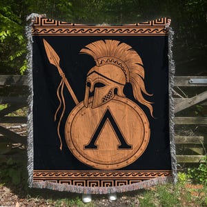 May include: A black woven throw blanket featuring a large orange Spartan helmet, a spear, and a circular shield with a prominent Lambda symbol. The design includes a Greek key border at the top and bottom, finished with decorative fringe edges.