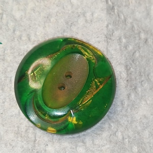 May include: A round, translucent green button featuring a recessed, oval-shaped center with two holes. The button has a smooth, glossy finish with subtle internal golden-yellow inclusions and a slightly darker, textured appearance in the center.