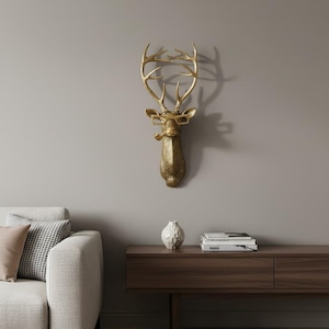 May include: A gold-coloured wall-mounted deer head sculpture featuring a textured finish. The deer is depicted wearing rectangular-framed glasses and holding a pipe in its mouth. The antlers are tall and branched, extending upwards from the head.
