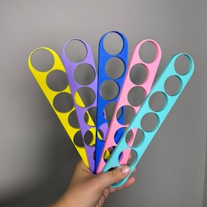May include: Five elongated, flat, plastic-like tools arranged in a fan shape. Each tool features four circular cutouts along its length. The colors of the tools from left to right are bright yellow, purple, royal blue, light pink, and turquoise.