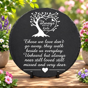 May include: A round black slate memorial plaque featuring a white engraved tree design with a heart-shaped canopy, a small bird, and the text "I AM always WITH YOU Kaila 1963-2025" followed by the quote "Those we love don't go away, they walk beside us everyday. Unheard but always near still loved still missed and very dear." with a small heart icon at the bottom.