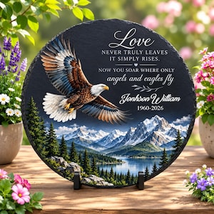 May include: A round black slate memorial plaque featuring a flying bald eagle above a mountain lake and pine forest. The text reads: Love NEVER TRULY LEAVES IT SIMPLY RISES. NOW YOU SOAR WHERE ONLY angels and eagles fly Jonhson William 1960-2026.