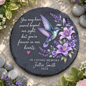 May include: A round black slate memorial plaque featuring a hummingbird, purple lilies, and the text: You may have soared beyond our sight, but you're forever in our hearts. IN LOVING MEMORY Jullia Smith 2026, with a small pink heart icon.