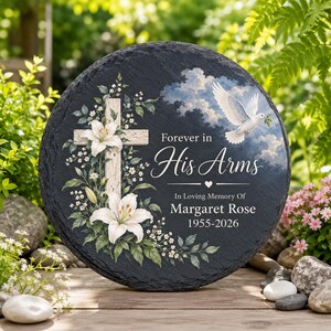 Op de afbeelding: Een ronde zwarte leistenen gedenkplaat met een wit kruis versierd met witte lelies, een vliegende witte duif tegen een bewolkte lucht, en de tekst Forever in His Arms, In Loving Memory of Margaret Rose, en de jaartallen 1955-2026.
