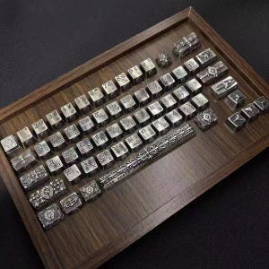 Puede incluir: Un conjunto de teclas de teclado en plata metálica con intrincados grabados en relieve de estilo gótico, símbolos y letras. La colección incluye una barra espaciadora larga con volutas detalladas y texto, dispuesta ordenadamente en una bandeja de madera oscura.