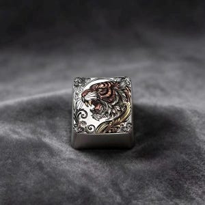 May include: A square mechanical keyboard keycap featuring a detailed, engraved illustration of a roaring tiger head. The design incorporates intricate scrollwork patterns in metallic tones of silver, bronze, and gold against a brushed metal surface.