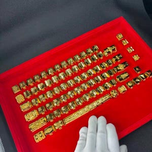Peut inclure: Une collection de touches de clavier en métal doré disposées en rangées sur un plateau de présentation rouge vif. L'ensemble comprend diverses touches carrées avec des lettres et symboles gravés, ainsi qu'une longue barre d'espace ornée.