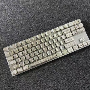 Könnte beinhalten: Eine mechanische Tastatur mit metallisch silbernem Finish und komplizierten, geprägten Blumen- und Ornamentmustern auf jeder Tastenkappe. Das Layout umfasst Standard-Alphanumerik-Tasten, eine gravierte Leertaste und den Text XTRFY.