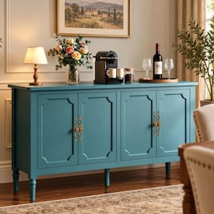 May include: A teal four-door sideboard cabinet with ornate gold-toned metal hardware. The top surface holds a table lamp, a glass vase with a floral arrangement, a black coffee machine with two mugs, a glass jar, and a wine bottle on a tray.