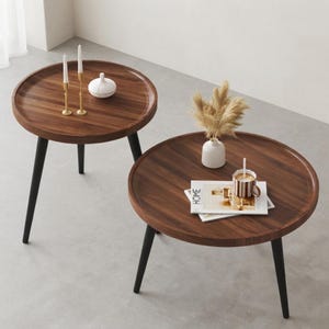 May include: Two round coffee tables with dark brown wood grain tops and raised edges, each supported by three slender black tapered legs. The tables are arranged with decorative items including two gold candlesticks, a white vase, and a mug.