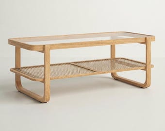 May include: A rectangular coffee table featuring a light-toned wood frame with rounded edges. The table includes a clear glass top surface and a lower shelf constructed from a fine-mesh metal grid, supported by two continuous U-shaped wooden legs.