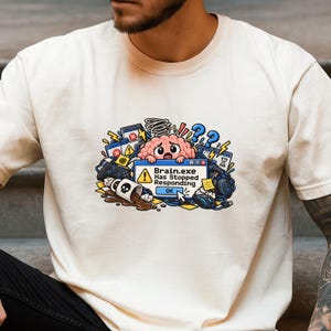 Può includere: Una t-shirt color crema con la grafica di un cervello rosa dei cartoni animati dietro una finestra di errore del computer con la scritta Brain.exe Has Stopped Responding. Il design include una tazza rovesciata, controller e cuffie.