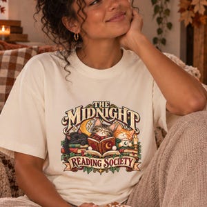Può includere: Una t-shirt color crema con una grafica di tre gatti che leggono libri e il testo "THE MIDNIGHT READING SOCIETY" in un carattere decorativo. L'illustrazione include un gatto nero, un gatto tigrato e un gatto arancione circondati da libri.