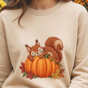 May include: A beige crewneck sweatshirt featuring a central embroidered graphic of a brown squirrel resting on top of a large orange pumpkin, surrounded by small red, green, and yellow autumn leaves and green vines with a textured stitch design.