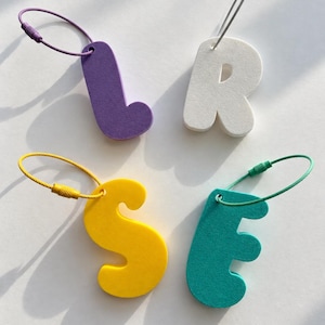May include: Four letter-shaped keychains in purple, white, yellow, and teal. Each letter features a rounded, bold font and is attached to a thin, circular metal wire cable loop with a screw-top closure matching the color of the letter.