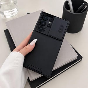 Könnte beinhalten: Eine mattschwarze Smartphone-Hülle mit einer verschiebbaren Abdeckung für das Kameraobjektiv. Die Hülle verfügt über einen rechteckigen Ausschnitt für das Kameramodul und eine glatte, einfarbige Rückseite mit Schiebemechanismus.