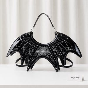 Puede incluir: Un bolso de mano de charol negro con forma de alas de murciélago y estampado de telaraña blanca. El bolso cuenta con ojales metálicos plateados en los bordes superiores y un asa. Una pequeña tarjeta blanca con el texto BagNotBag está colocada.