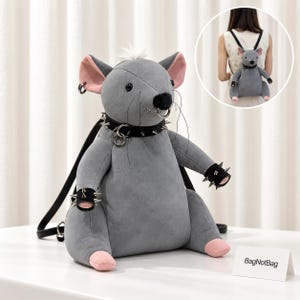 May include: A grey plush rat-shaped backpack featuring a black collar with metal spikes, a nose ring, and matching spiked wrist cuffs. The backpack has pink inner ears and feet, black shoulder straps, and a small white tuft of hair on its head.