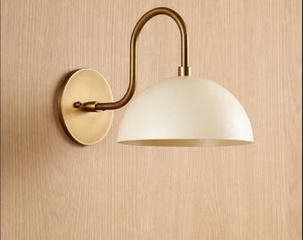 May include: A wall-mounted lamp featuring a circular brass backplate and a curved brass arm that supports a matte off-white dome-shaped shade. The fixture has a clean, metallic finish on the hardware and a smooth, solid-coloured light diffuser.