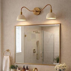 May include: A two-light wall sconce with a brushed gold finish and cream-coloured dome shades mounted above a rectangular mirror with a gold frame. The fixture features curved arms extending from a central circular wall plate.