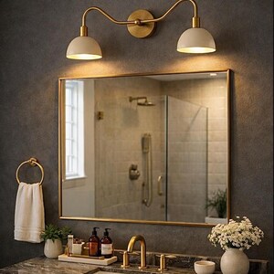 May include: A bathroom vanity featuring a rectangular mirror with a gold frame, a double-light wall sconce with cream shades, a gold faucet, a white towel on a gold ring, and a marble countertop with a white sink and decorative accessories.