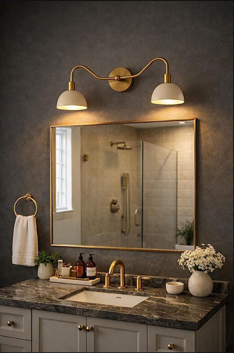 May include: A bathroom vanity featuring a rectangular mirror with a gold frame, a double-light wall sconce with cream shades, a gold faucet, a white towel on a gold ring, and a marble countertop with a white sink and decorative accessories.