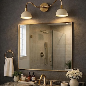 May include: A bathroom vanity set featuring a rectangular mirror with a gold frame, a double-light wall sconce with white dome shades and gold hardware, a gold tap, a marble countertop, and a gold towel ring holding a white hand towel.