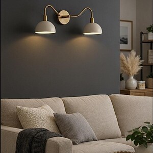 May include: A double wall light featuring a brass-toned metal frame with two curved arms extending from a central circular wall mount. The fixture includes two dome-shaped off-white shades that direct light downwards, creating a warm glow.