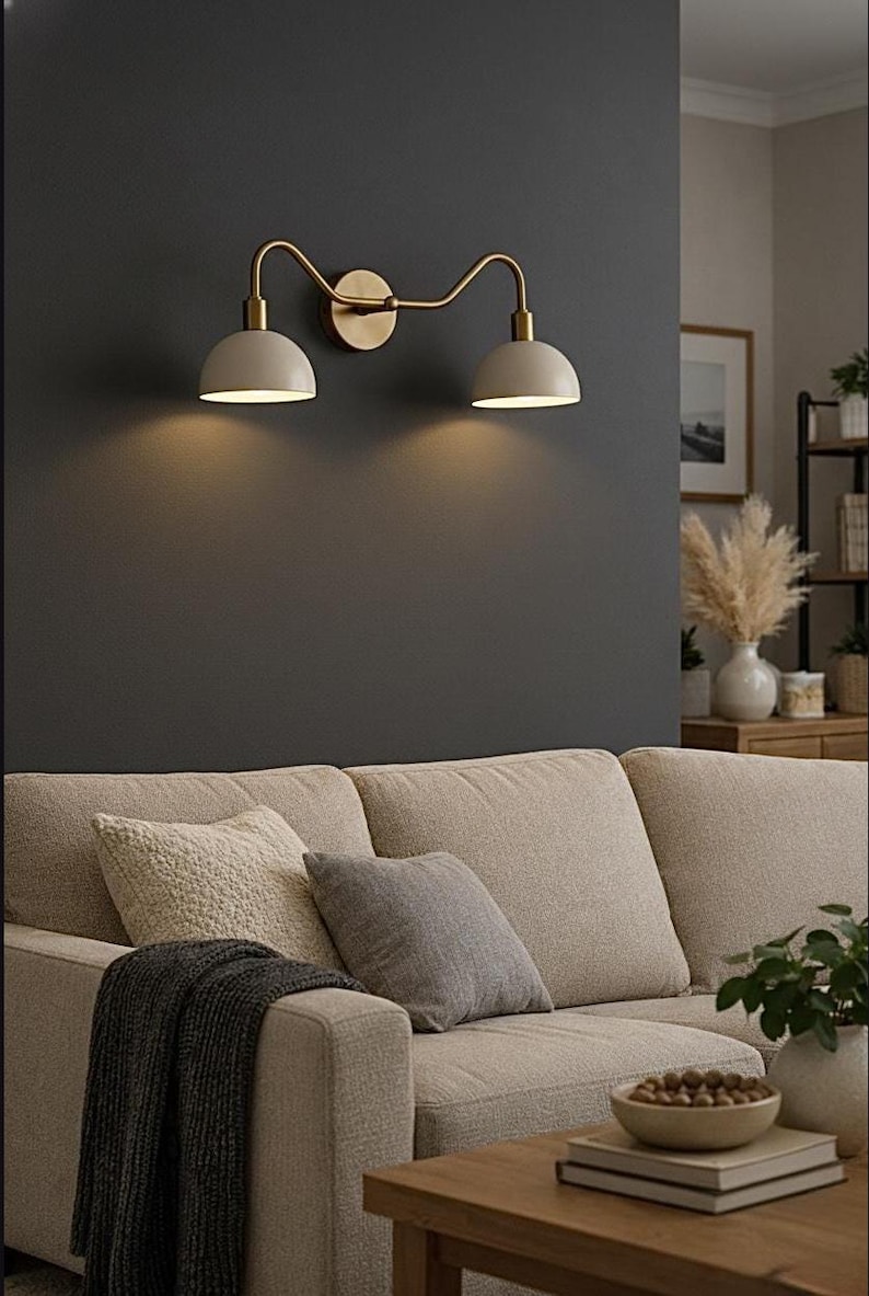 May include: A double wall light featuring a brass-toned metal frame with two curved arms extending from a central circular wall mount. The fixture includes two dome-shaped off-white shades that direct light downwards, creating a warm glow.