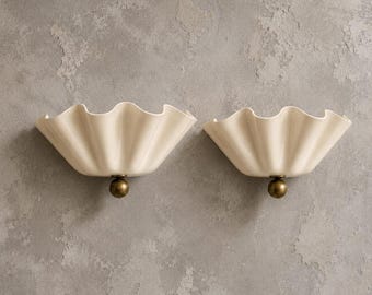 May include: A pair of wall lights featuring off-white, ruffled, fan-shaped shades made of a smooth material. Each shade is attached to a circular, metallic, antique brass-toned base that connects to the wall, creating a symmetrical lighting design.