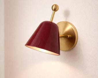 May include: A wall-mounted lamp featuring a glossy, deep red, bell-shaped shade attached to a brushed brass circular base. A thin brass arm extends from the base, connecting to the top of the shade, which is topped with a small brass sphere.