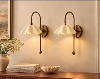 May include: A pair of wall-mounted lamps featuring curved brass-toned metal arms and off-white, scalloped, bell-shaped shades. Each lamp has a circular wall plate and a downward-facing light source that emits a warm, soft glow.