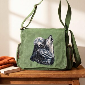 Puede incluir: Un bolso tipo mensajero color verde salvia con un estampado gráfico de un lobo aullando a la luna llena sobre una cordillera, un bosque de pinos y un lago. El bolso cuenta con una correa ajustable, una solapa frontal y bolsillos laterales con cremalleras.