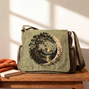 May include: An olive green canvas messenger bag featuring a circular graphic print on the front flap. The design depicts a large crescent moon framing a landscape with a gnarled tree, a mountain range, a lake, and a full moon in the night sky.