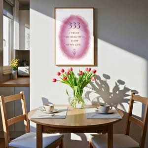 Puede incluir: Un cuadro decorativo enmarcado con un fondo ovalado de acuarela rosa que incluye el texto 333 I TRUST THE BEAUTIFUL FLOW OF MY LIFE y una pequeña ilustración de un tulipán. El cuadro cuelga sobre una mesa redonda de madera con un florero.