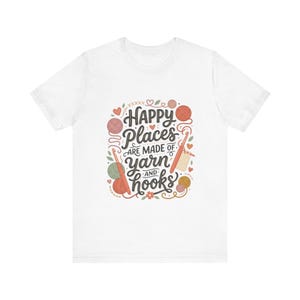May include: A white short-sleeved t-shirt featuring a central graphic with the text "Happy Places ARE MADE OF yarn AND hooks". The design includes illustrations of colourful yarn balls, crochet hooks, small leaves, and hearts in shades of red, pink, and green.