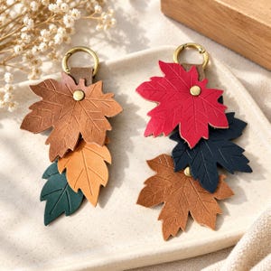 Può includere: Due portachiavi con foglie d'acero in pelle sovrapposte nei colori autunnali. Un portachiavi presenta foglie marroni, ocra e verde scuro; l'altro foglie rosse, blu scuro e ocra. Entrambi includono un anello in metallo dorato e un rivetto.