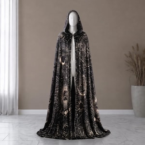 May include: A floor-length hooded cloak in black featuring a gold celestial print of suns, moons, stars, and astrological constellations. The garment has a pointed hood and a full-length open front, draping down to the floor in a wide, flowing silhouette.
