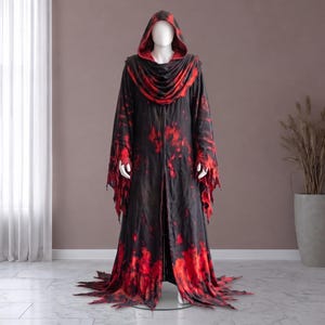 May include: A floor-length hooded robe featuring a black base with a vibrant red tie-dye pattern. The garment includes long, wide sleeves with jagged, distressed edges and a matching frayed hemline that pools at the base of the mannequin.